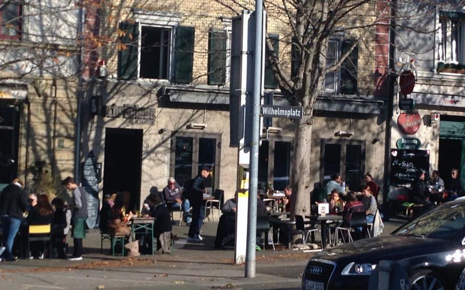 The lunch-time crowd gathers on a warm November afternoon outside Schraeglage Meals and More, where there is indoor and outdoor seating. The eatery is located along Wilhelmsplatz, a street in Stuttgart, Germany, that boasts several popular restaurants.