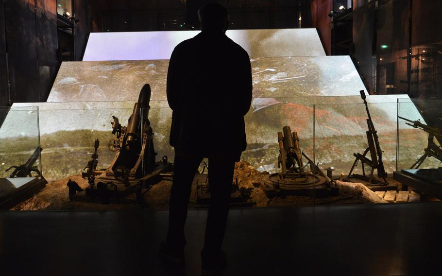 A visitor stands in front of the battlefield display at the Verdun Memorial Museum. In the foreground are various weapons used, while videos and photos projected on the large screens show explosions, soldiers crawling through the trenches and the aftermath. Visitors can put on headphones to  listen to the sounds of the carnage.