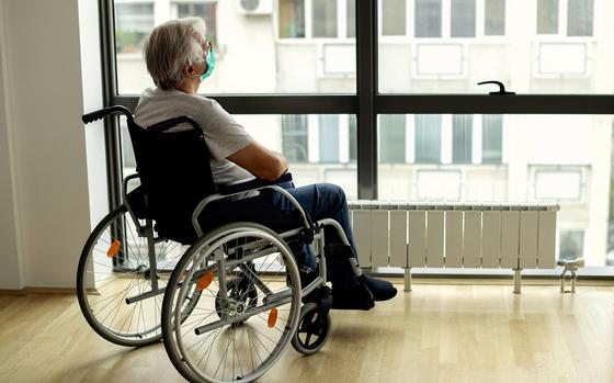 Pensive mature man in wheelchair wearing protective face mask while looking through the window during home quarantine.